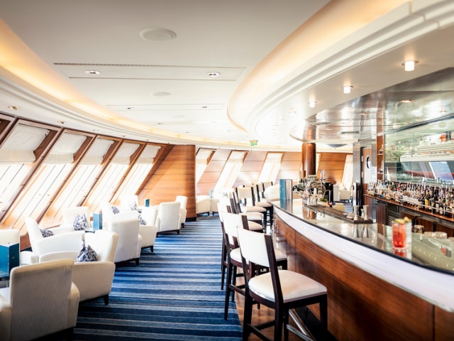 Queen Mary 2's Commodore Club, showing curved wooden bar and comfortable seating near front-facing windows