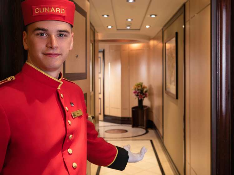 A bellhop in Cunard uniform holds open the door to a Queens Grill Suite on Queen Mary 2