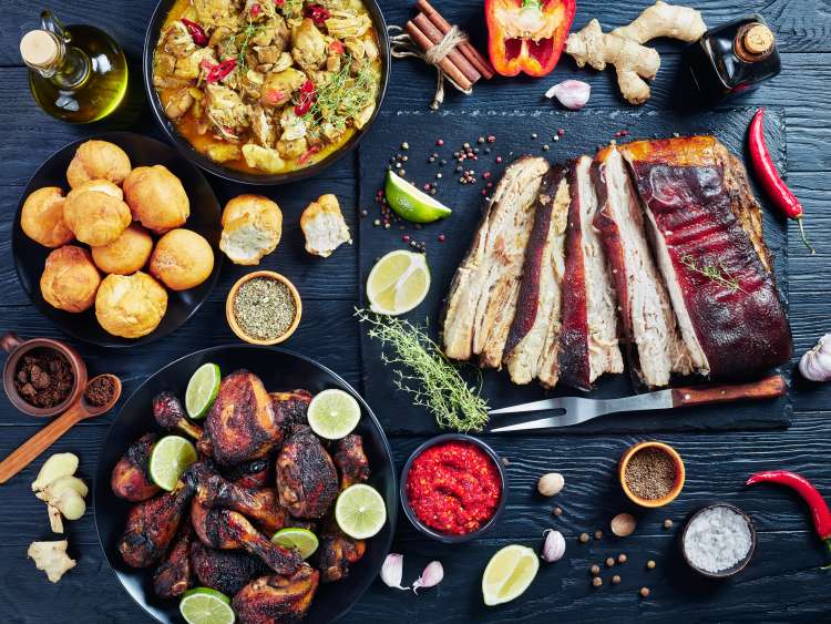 set of caribbean dishes, jerk pork belly, chicken curry, fried dumplings, roasted chicken thighs and drumsticks on plates on a black wooden table with spices and herbs, view from above, flat lay; Shutterstock ID 1476457733; purchase_order: -; job: -; client: -; other: -