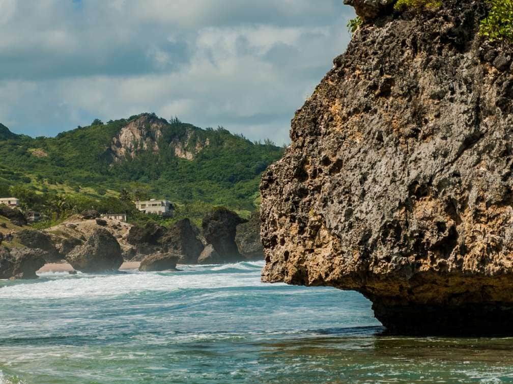 Coastline of Barbados featuring a large rock in the water in the foreground with an outline of green trees in the background