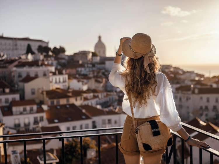 Blonde woman standing on the balcony and looking at coast view of the southern european city with sea during the sunset, wearing hat, cork bag, safari shorts and white shirt; Shutterstock ID 1398214628; Purchase Order: -