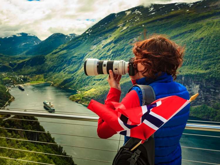 Tourism holidays picture and traveling. Woman tourist enjoying fjord landscape Geirangerfjord from Ornesvingen eagle road viewpoint, taking photo with camera, Norway.