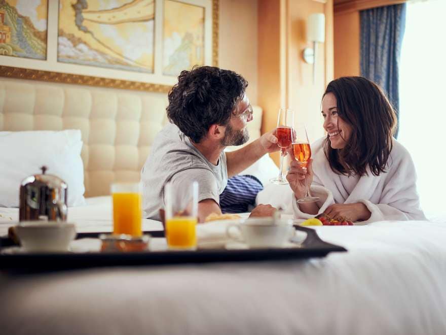 A couple lie on a bed in comfortable robes and clink glasses of champagne over breakfast