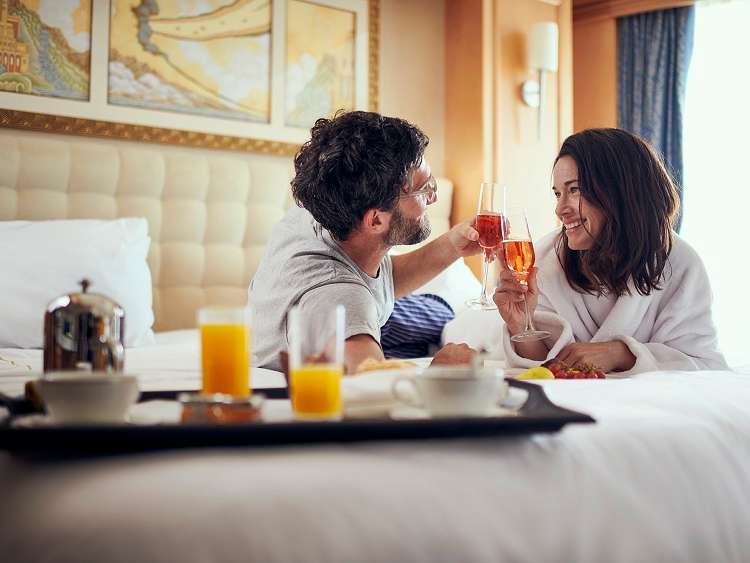 A couple lie on a bed in comfortable robes and clink glasses of champagne over breakfast