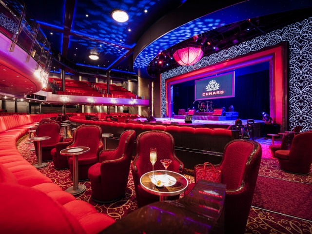 The Royal Court Theatre on Cunard's Queen Mary 2, showing a lit up stage set up for a presentation, rows of plush seating, and cabaret-style tables and chairs