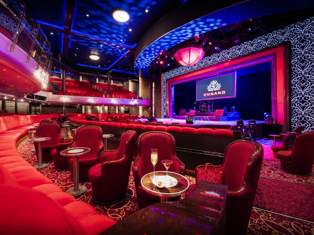 The Royal Court Theatre on Cunard's Queen Mary 2, showing a lit up stage set up for a presentation, rows of plush seating, and cabaret-style tables and chairs