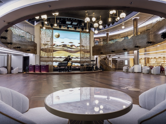 Queens Room on Queen Anne, showing dancefloor, upper deck mezzanine, and stage with ocean-themed backdrop and grand piano