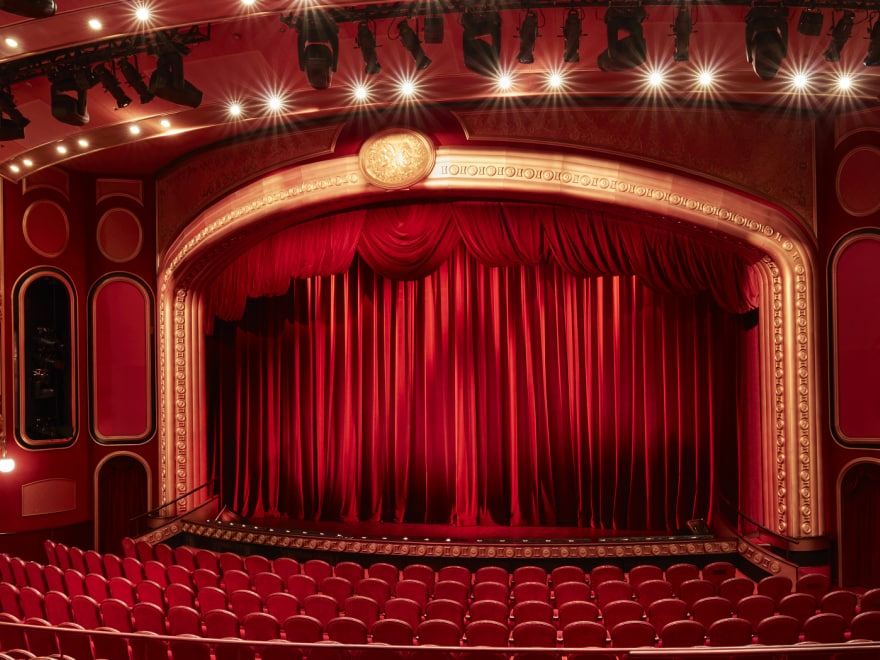 vThe Royal Court Theatre on Cunard's Queen Victoria, showing a curtained stage, rows of traditional theatre-style seating and boxes high up to the sides of the stage