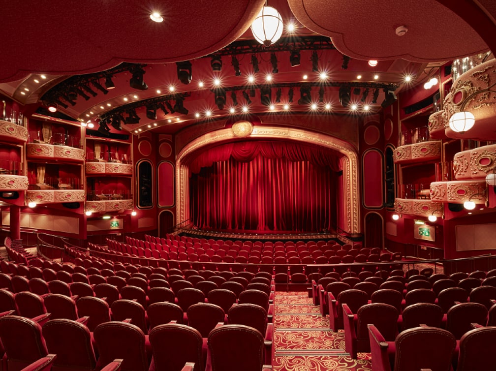 The Royal Court Theatre on Cunard's Queen Victoria, showing a curtained stage, rows of traditional theatre-style seating and boxes high up to the sides of the stage