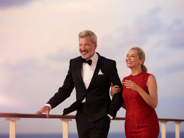 A middle-aged man and woman in formal dress walk, arms linked, along the deck of Queen Mary 2