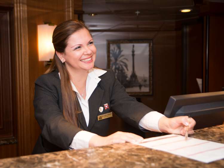 A smiling female Cunard crew member works at the on board reception desk 