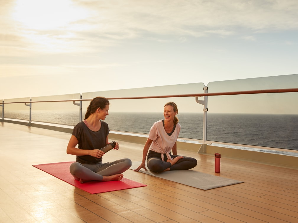 Two women sit on yoga mats on deck at sunset